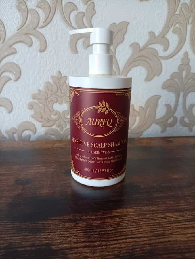 Aureo sensitive scalp shampoo bottle on a wooden surface with decorative wallpaper in the background
