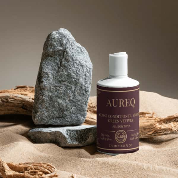 Gloss Conditioner, Smoky Green Vetiver bottle surrounded by rocks and sand, ideal for silky and manageable hair.