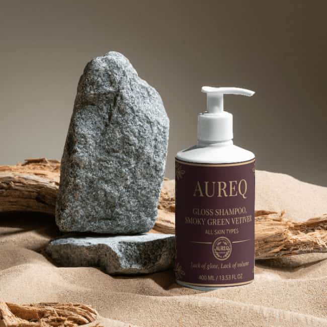 Gloss Shampoo, Smoky Green Vetiver bottle next to a stone on sandy surface, showcasing natural beauty.