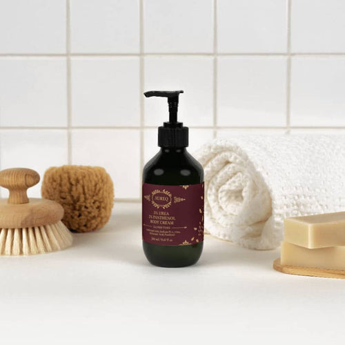 5% Urea + 2% Panthenol Body Cream bottle on spa table with towels and loofah.