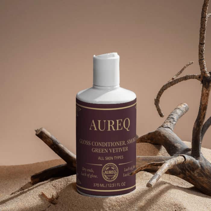 Aureq Gloss Conditioner, Smoky Green Vetiver bottle on a sandy surface with rustic branches.