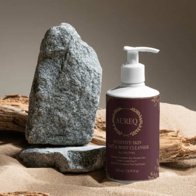 Sensitive Skin Face & Body Cleanser bottle next to stones on sandy background, highlighting gentle skincare.